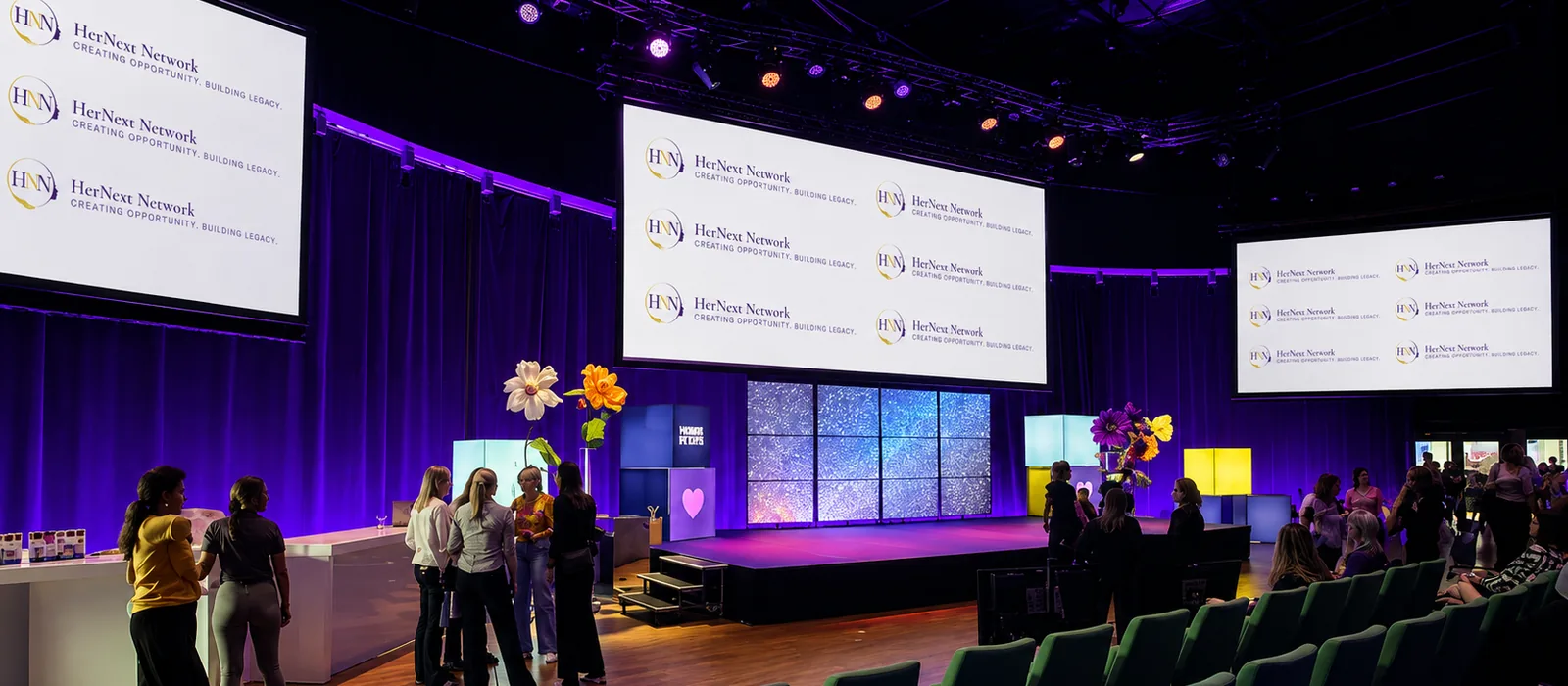 Delegates gathering in the hall at the HerNext Partnership Forum, the branded screens above the stage