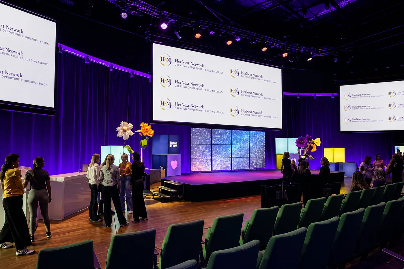 The stage and branded screens at a HerNext Network event, guests arriving