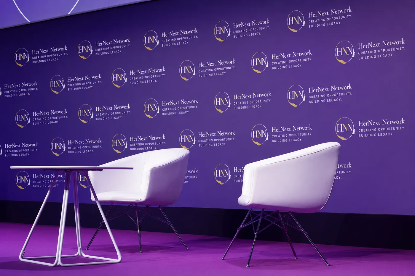 Two chairs set for a conversation in front of a HerNext Network media wall