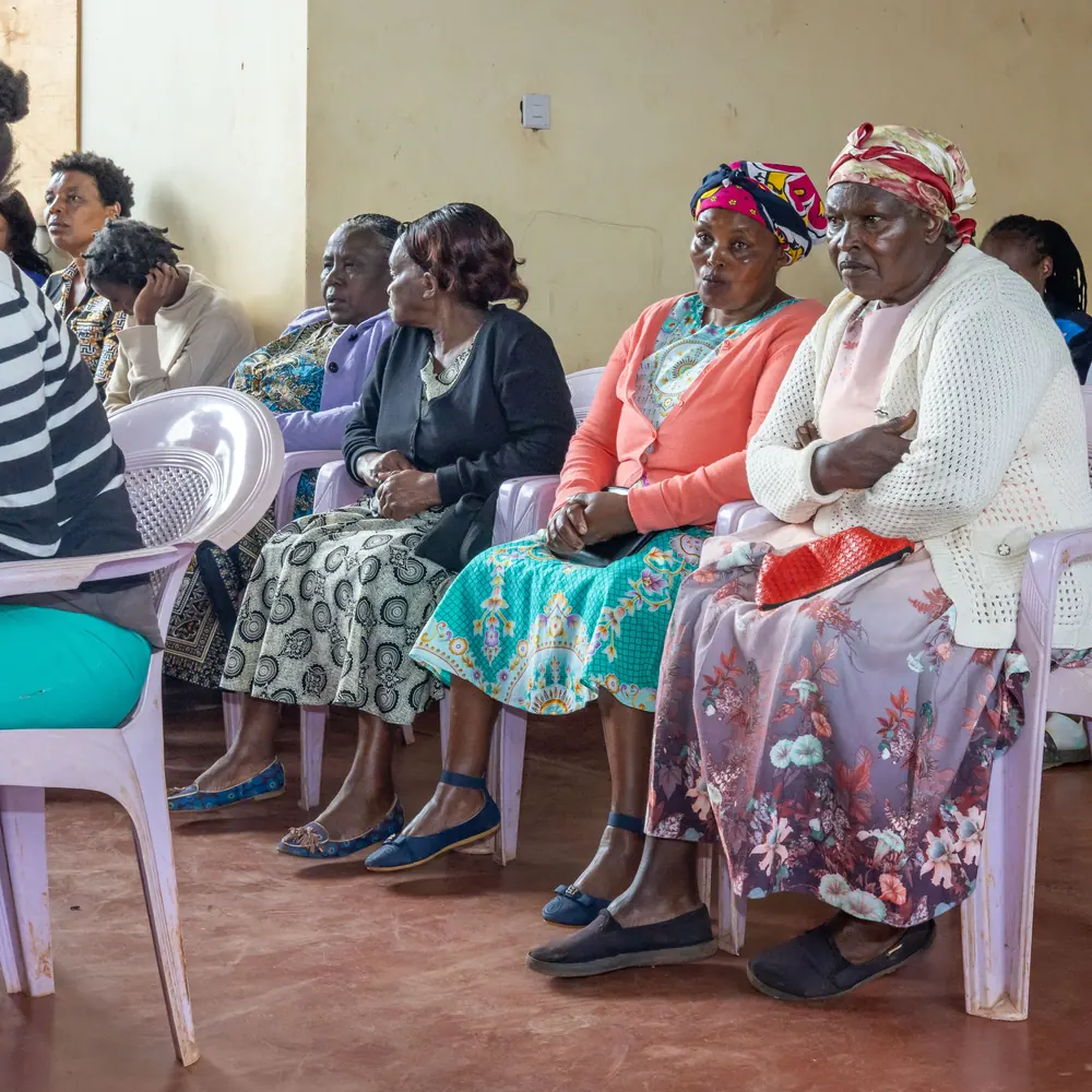 A row of older women in cardigans and head wraps seated on plastic chairs, listening